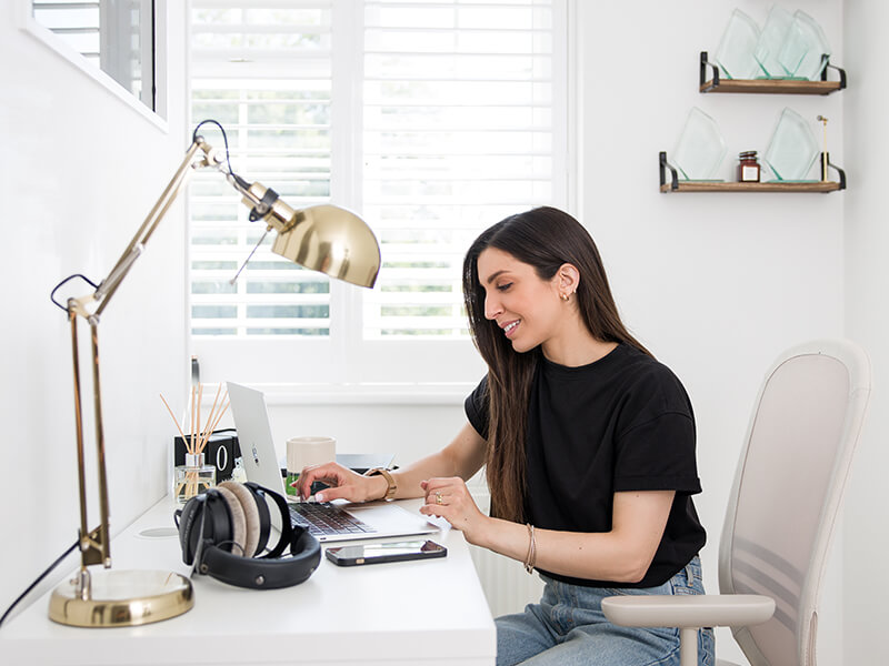 Alexia Kombou sat working in her home office. She is wearing blue jeans and a black t-shirt. Room colours are minimal, pale with a gold lamp and her voiceover awards on shelves in the background.