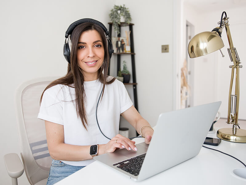 Alexia Kombou sat working in her home office. She is wearing headphones, blue jeans and a white t-shirt while typing at her laptop. She faces the camera, smiling.