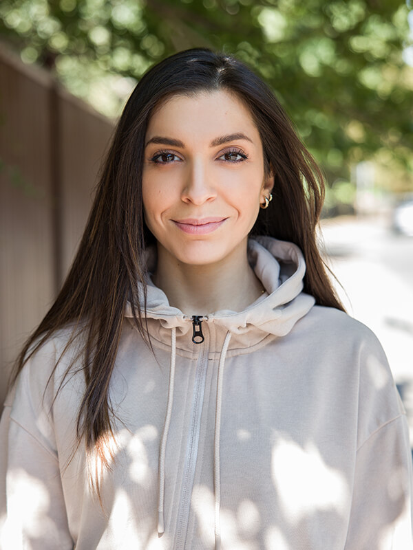 Female voiceover Alexia Kombou wearing a pale hoodie, smiling at the camera with out-of-foccus trees in the background.